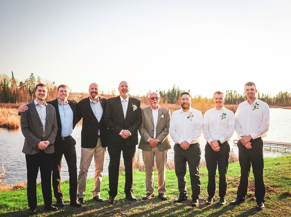 A group of well-dressed men in suits, blazers and dress shirts stand together outdoors by a waterfront at sunset.