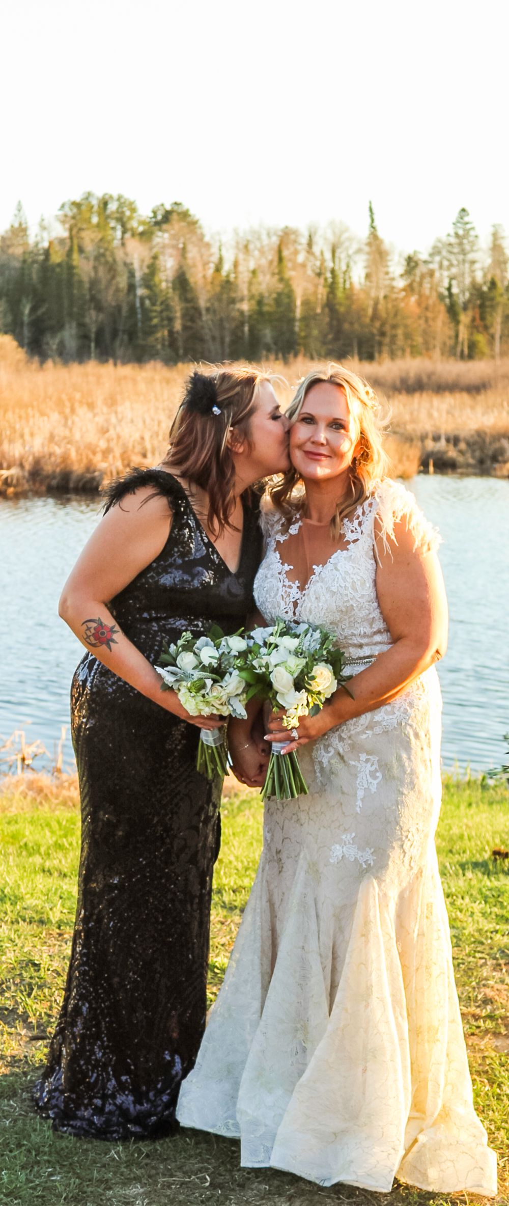 A wedding scene at sunset by a lake with autumn trees, featuring two people in formal dresses holding white bouquets.