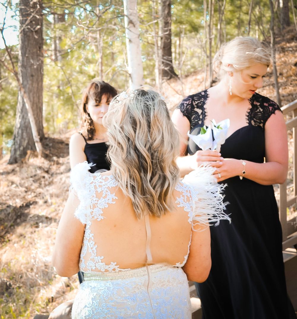 Three people in black and white formal attire gather outdoors in a sunlit wooded setting during a special occasion.