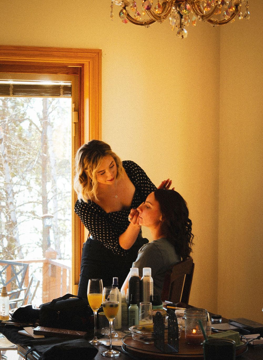 A makeup artist applies cosmetics at a vanity table with beverages and beauty products visible in a warmly lit room.