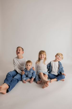 siblings in matching denim and white outfits sitting against a white backdrop laughing and being silly together for a family photoshoot