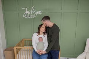A couple stands together in a nursery holding their newborn baby boy wrapped in a green swaddle in their macomb michigan home
