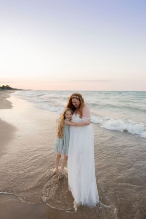 A pregnant mother in a white gown and daughter in blue dress embrace on a sandy beach at sunset in port huron michigan