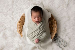 Newborn baby swaddled in sage green wrap sleeping peacefully in a wicker basket on a white fur rug.