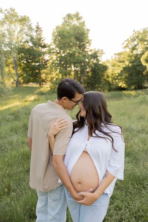 pregnant couple sharing a tender moment outdoors, woman cradling pregnant belly in a sunlit green field in Rochester michigan