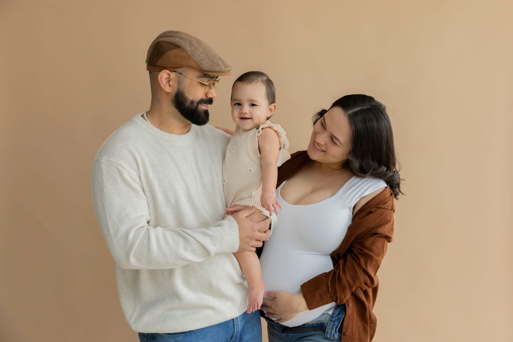 A pregnant family of three wearing casual clothing poses together against a neutral beige background at a studio in macomb michigan