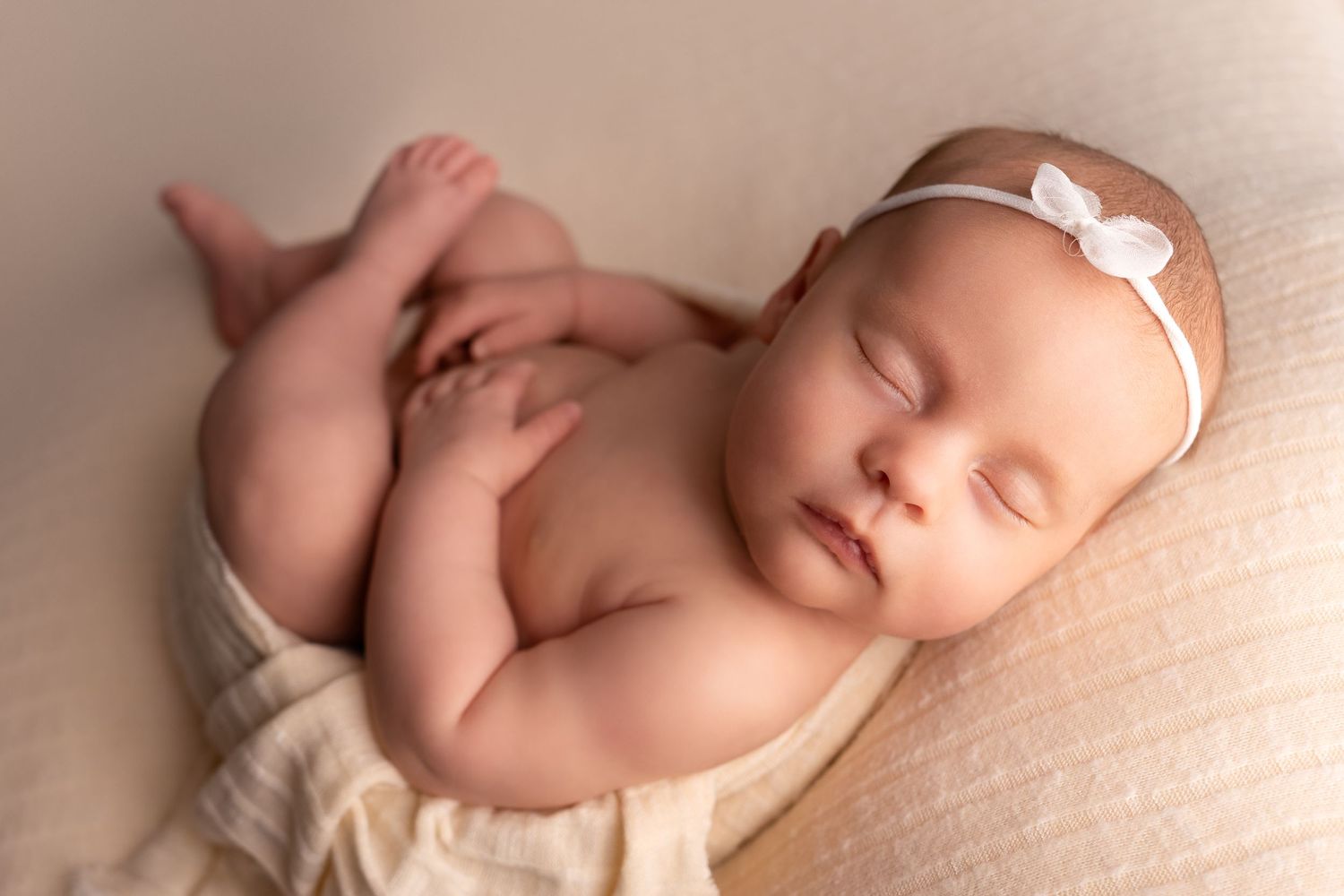 Sleeping newborn wearing white bow headband on cream colored blanket.