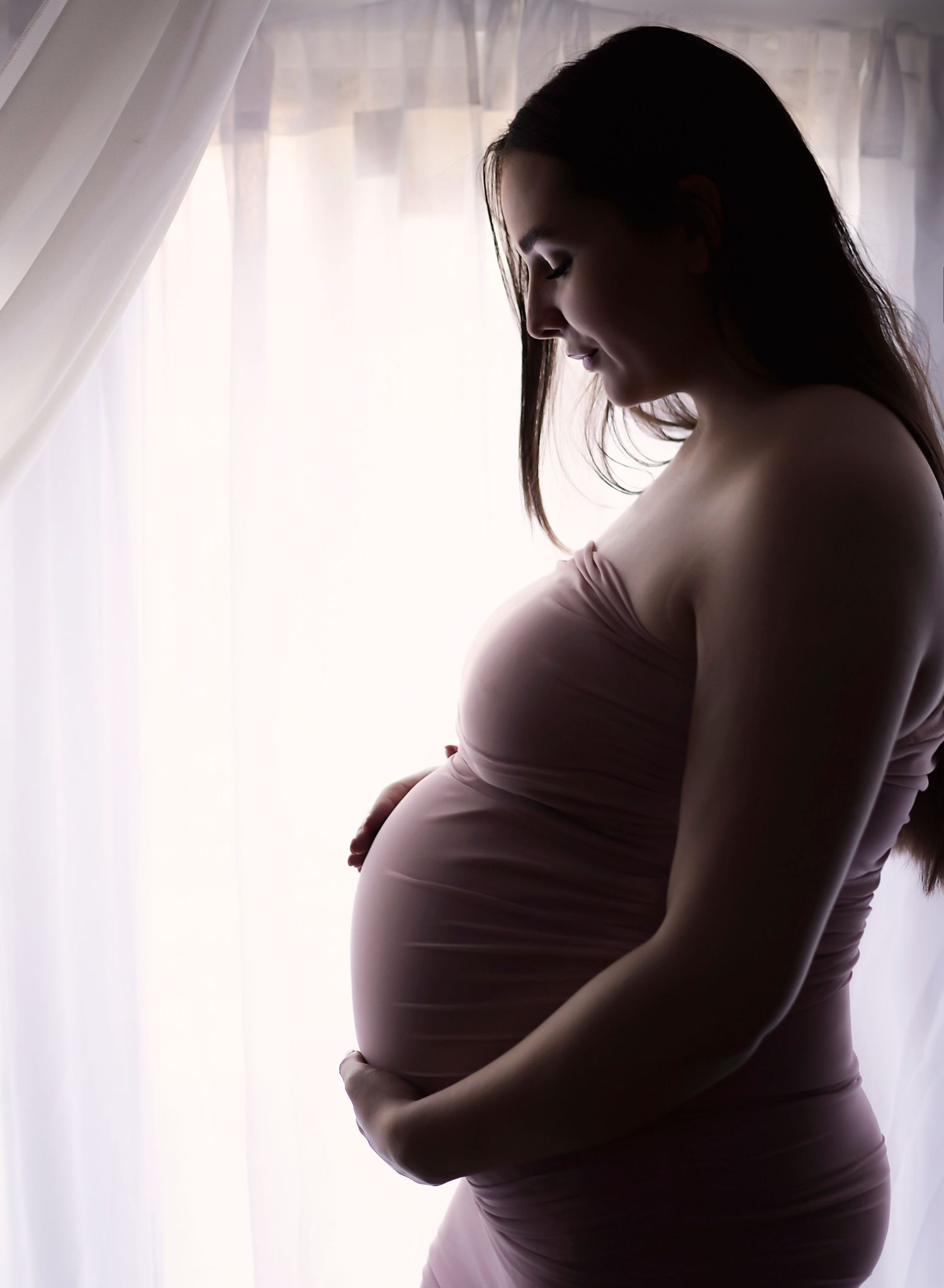Mother to be with her hands on her pregnant stomach, a silhouette shot with the natural light on a curtain backdrop.