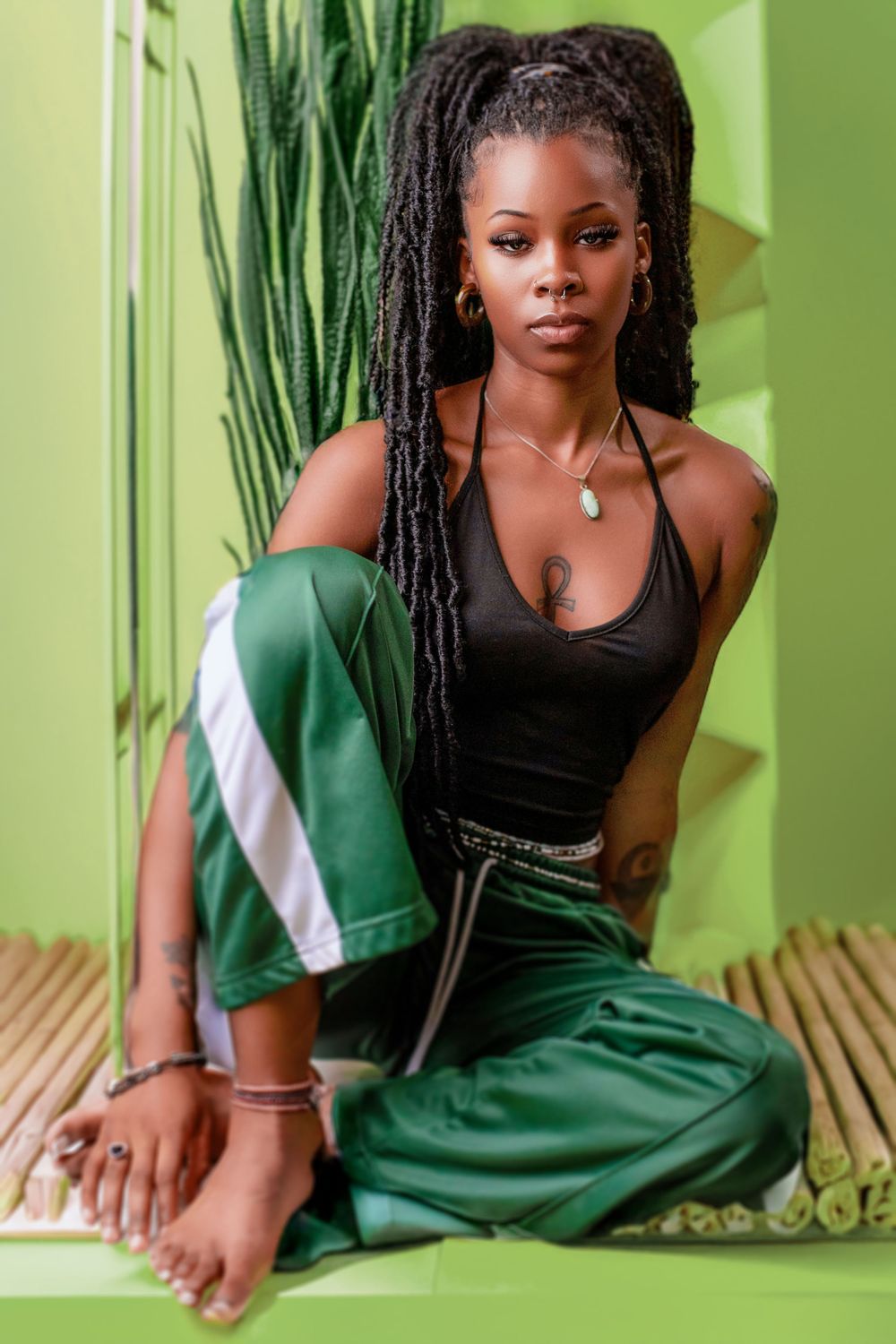 Young Black woman with long dreadlocks sits barefoot in green track pants and black top against a green background.