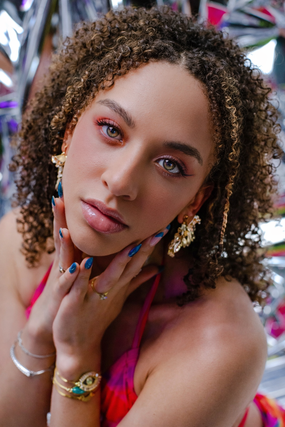 A person with curly hair and colorful makeup poses in multiple close-up shots wearing pink clothing.