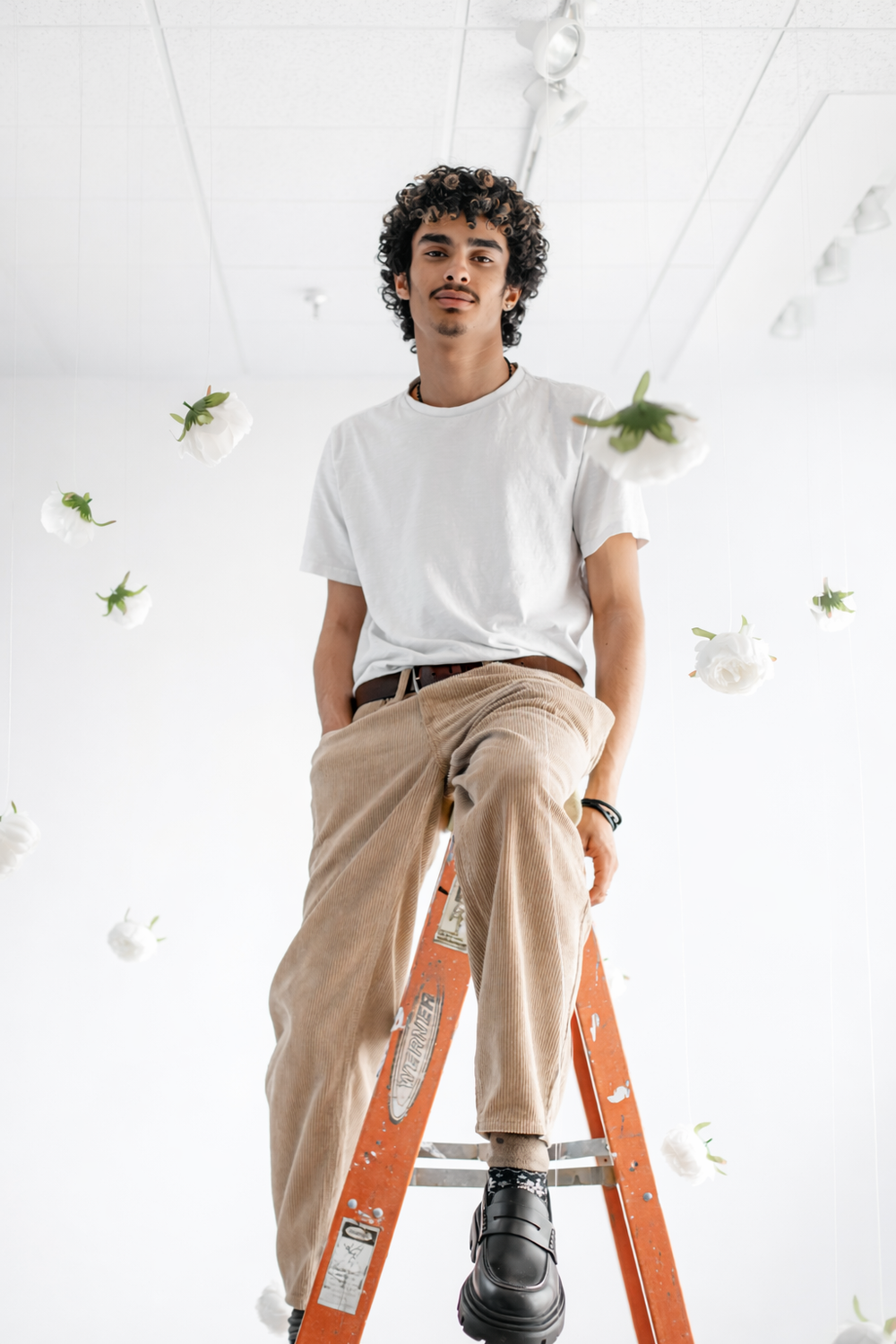 A person in white t-shirt and khaki pants sits on an orange ladder against a white background with floating green leaves.