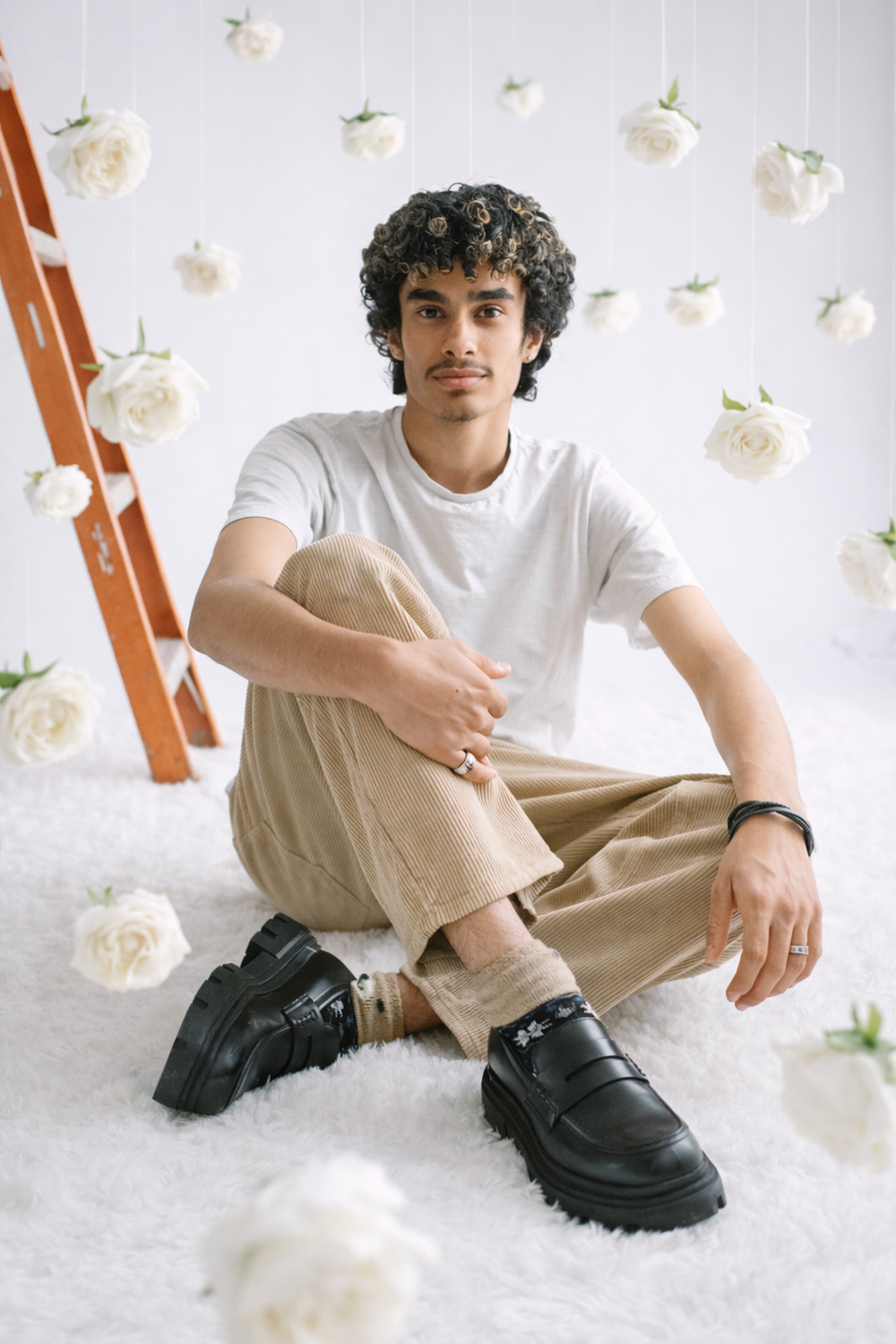 A person in a white t-shirt and khaki pants sits on the floor surrounded by hanging white roses and wearing chunky black boots.
