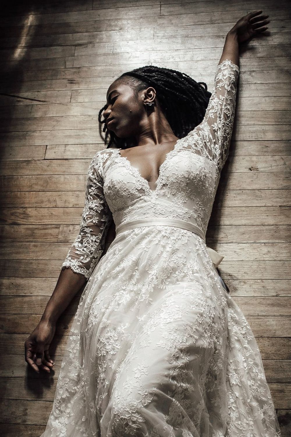 A Black woman in an elegant white lace wedding gown poses gracefully on a wooden floor with one arm raised.