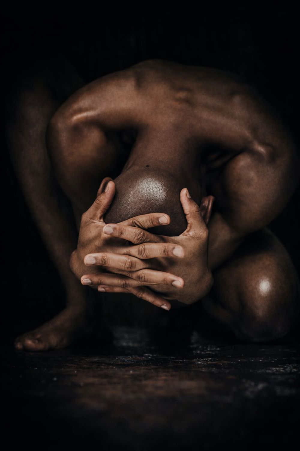 A muscular person crouched in a dark studio, hugging their knees in a powerful, emotive fine art pose.
