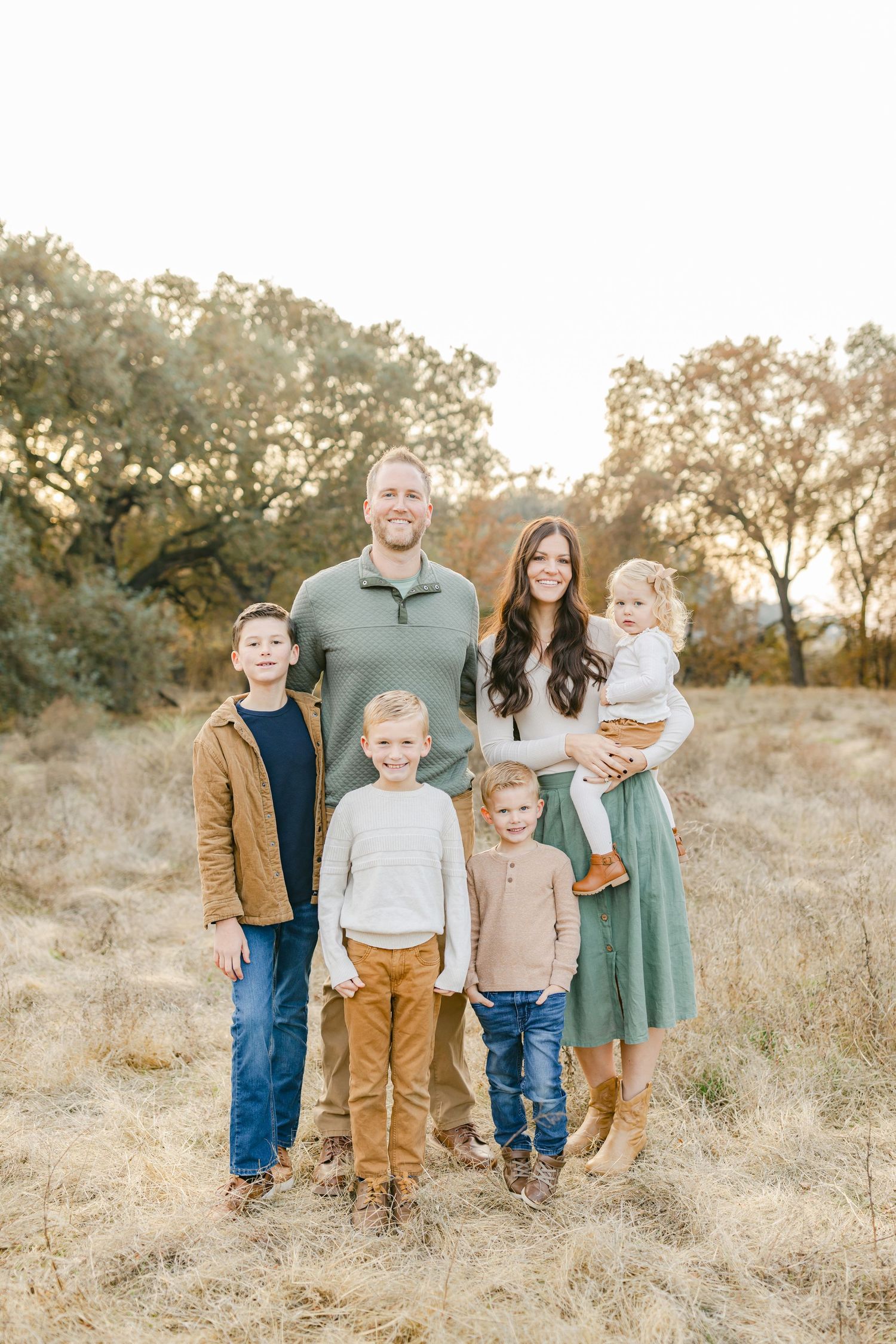 Family of six posing outdoors in a golden field, wearing coordinated fall outfits, with trees and warm sunset light behind them.