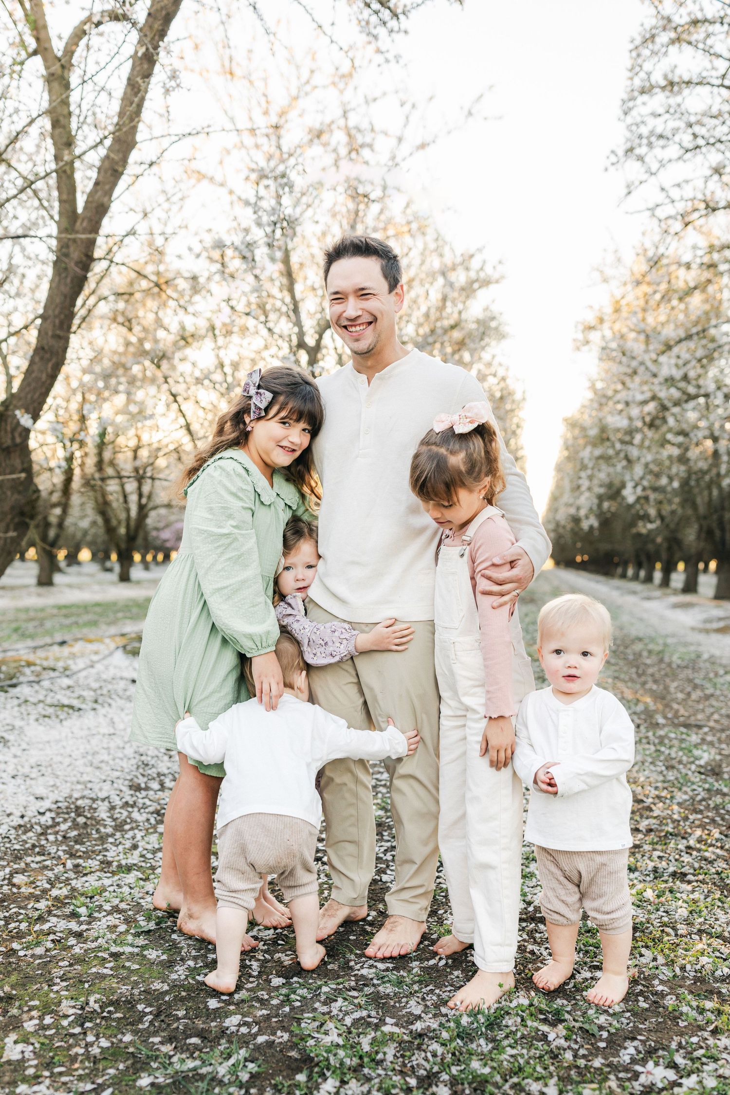 Family of six poses outdoors among blossoming trees, parents smiling with four young children in soft neutral and pastel tones.