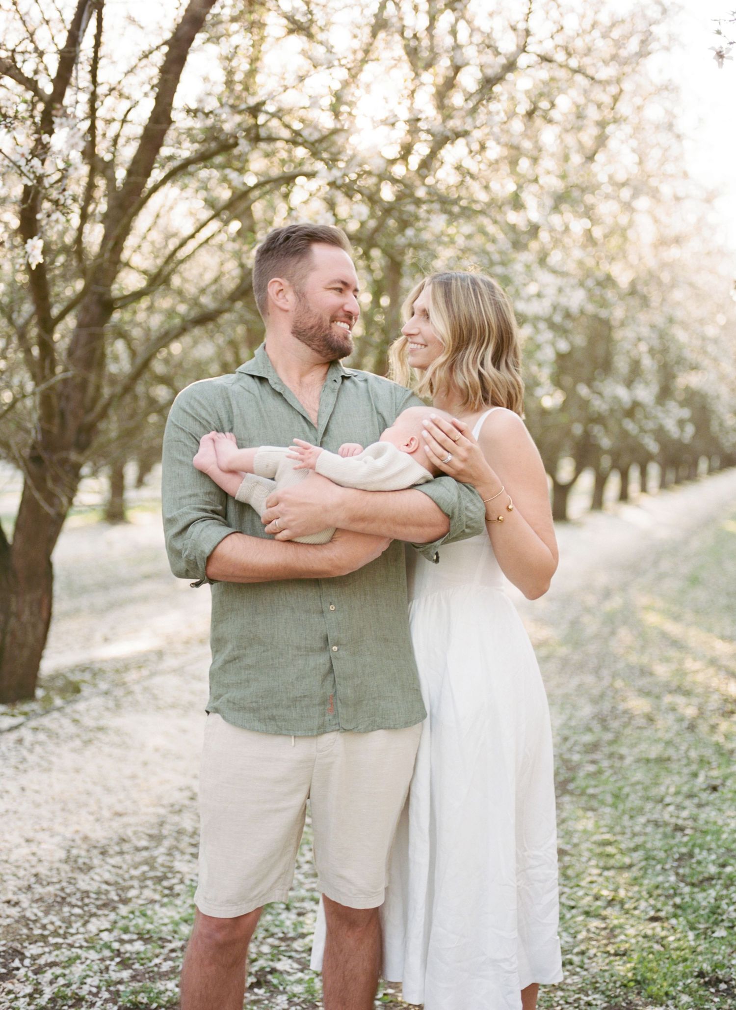 A couple smiles at each other while holding their newborn baby in a sunlit orchard with blooming trees.