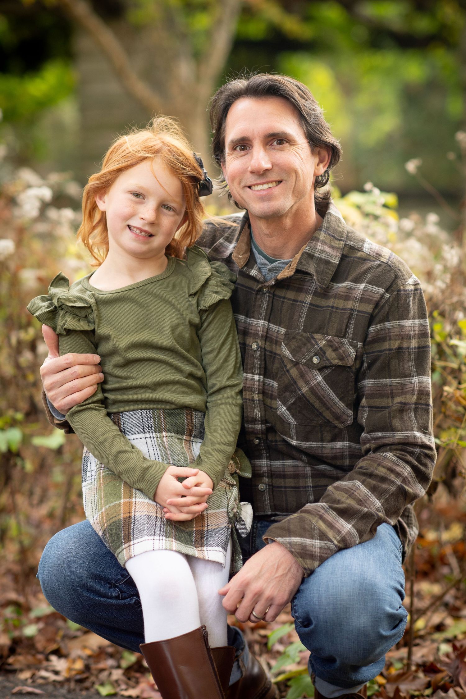 A smiling man in a plaid shirt crouches beside a red-haired girl in a green dress outdoors in autumn.