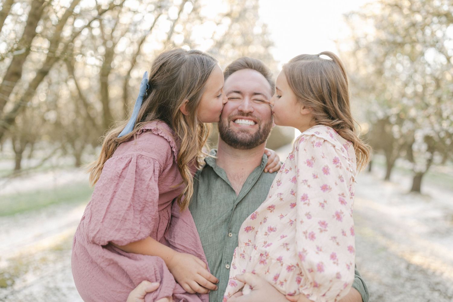 A bearded man smiles joyfully as two young girls in floral dresses kiss his cheeks outdoors.