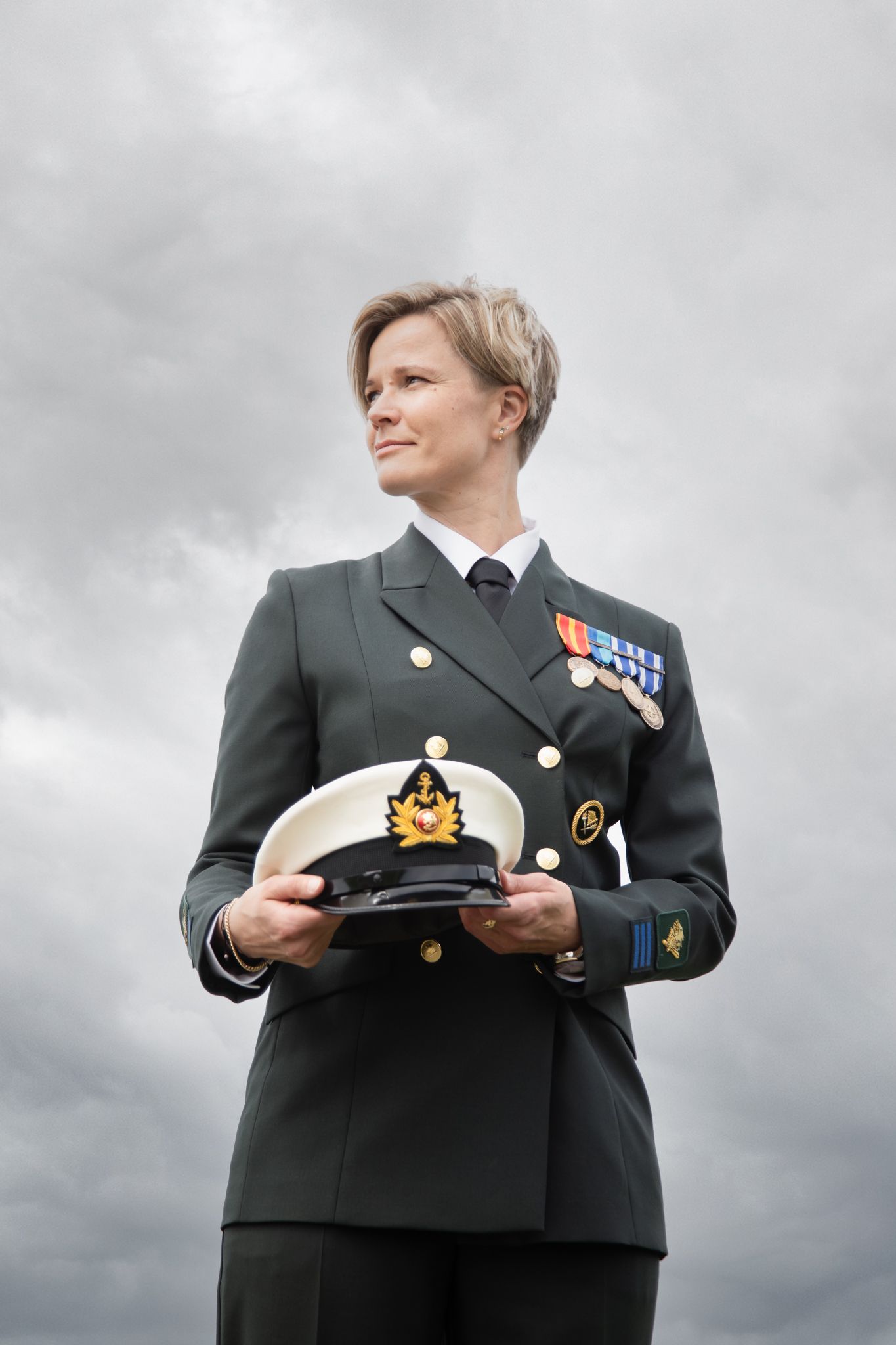 Female military officer in dress uniform holding a cap with medals on her chest against cloudy sky.