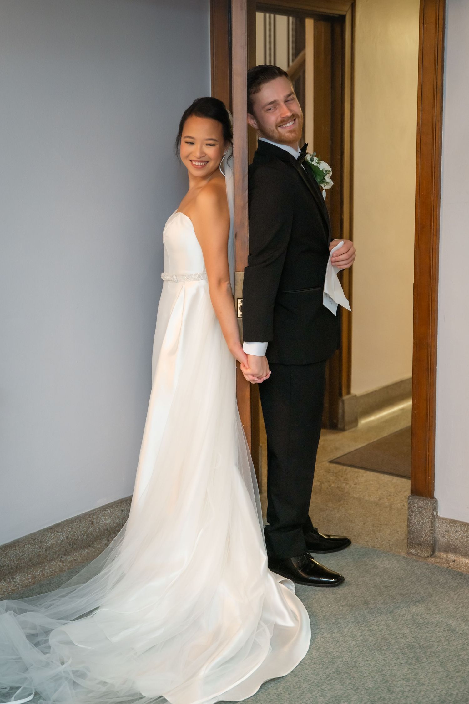 Bride in white gown and groom in black tuxedo hold hands, smiling, standing in a doorway on their wedding day.