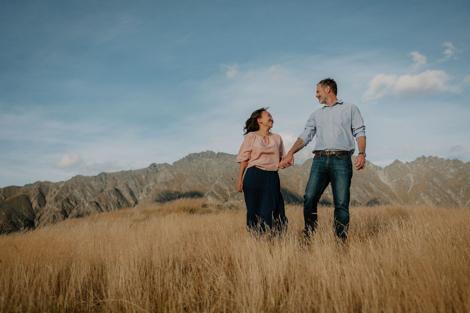 Couple holding hands in golden grass field with mountain backdrop under blue sky.