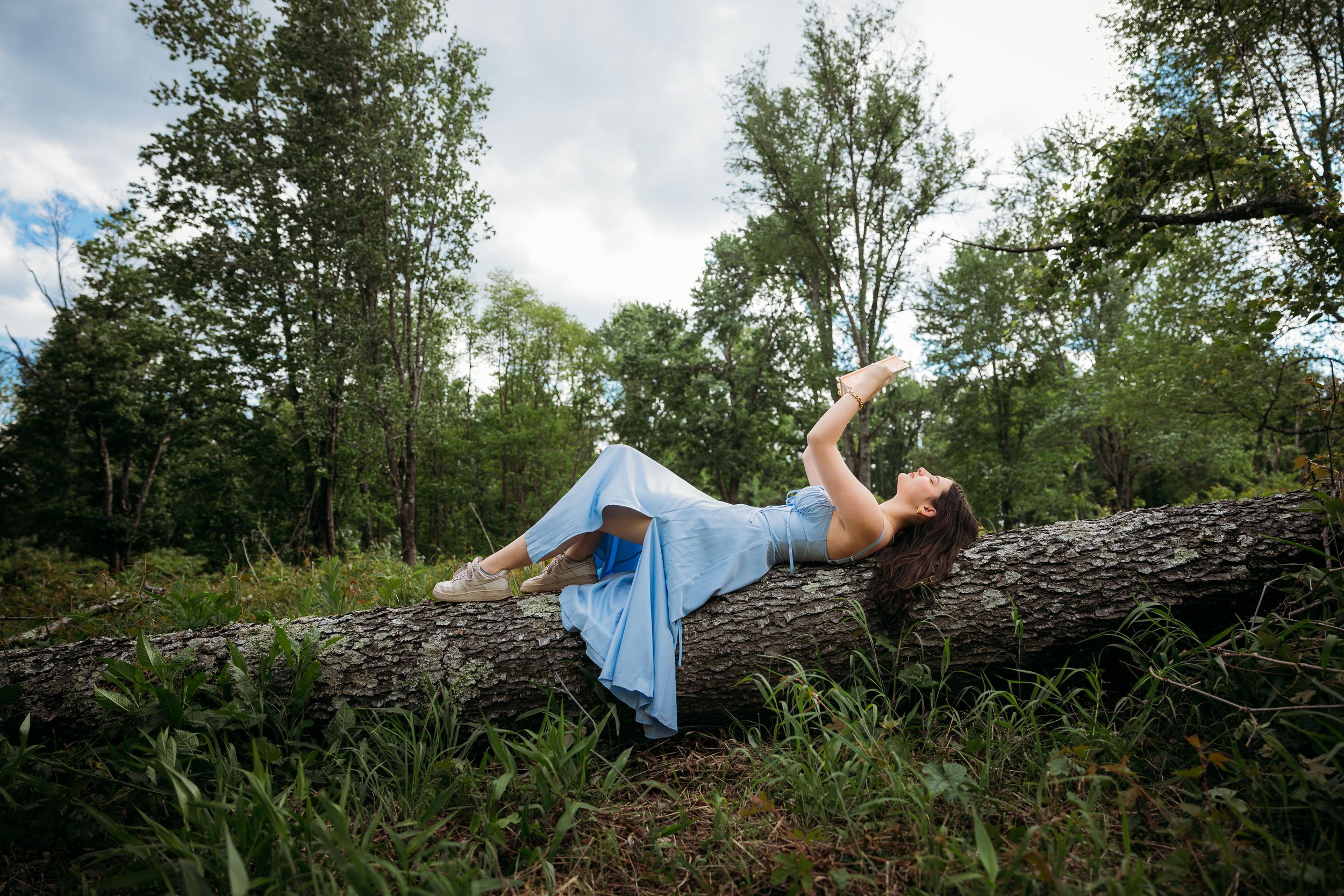 Woman in blue dress lying gracefully on a fallen log in a lush green forest under a cloudy sky.