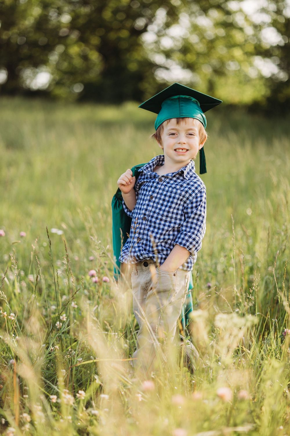 Young boy in green graduation cap and gown smiling in a sunny meadow field.