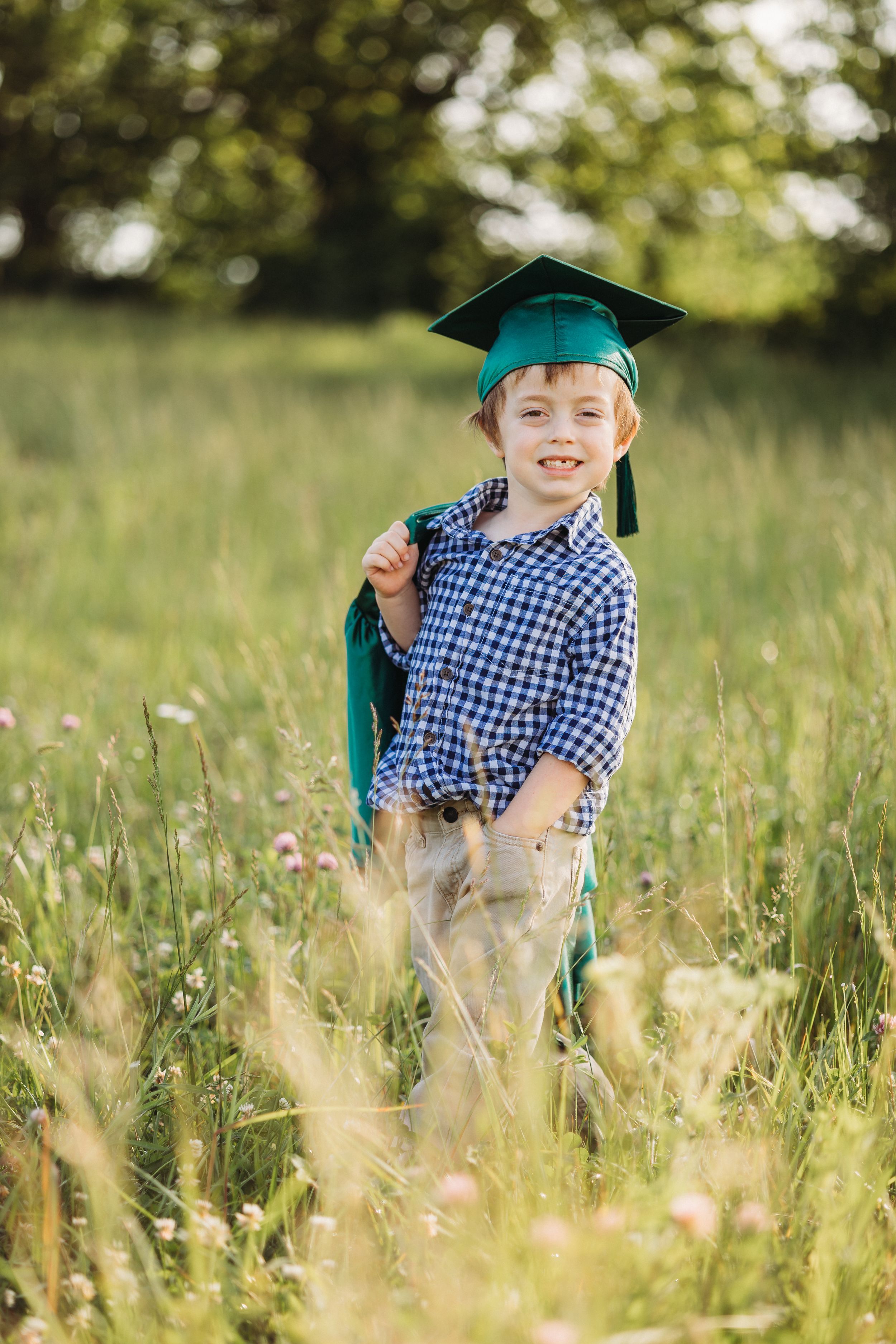 Young boy in green graduation cap and gown smiling in a sunny meadow field.