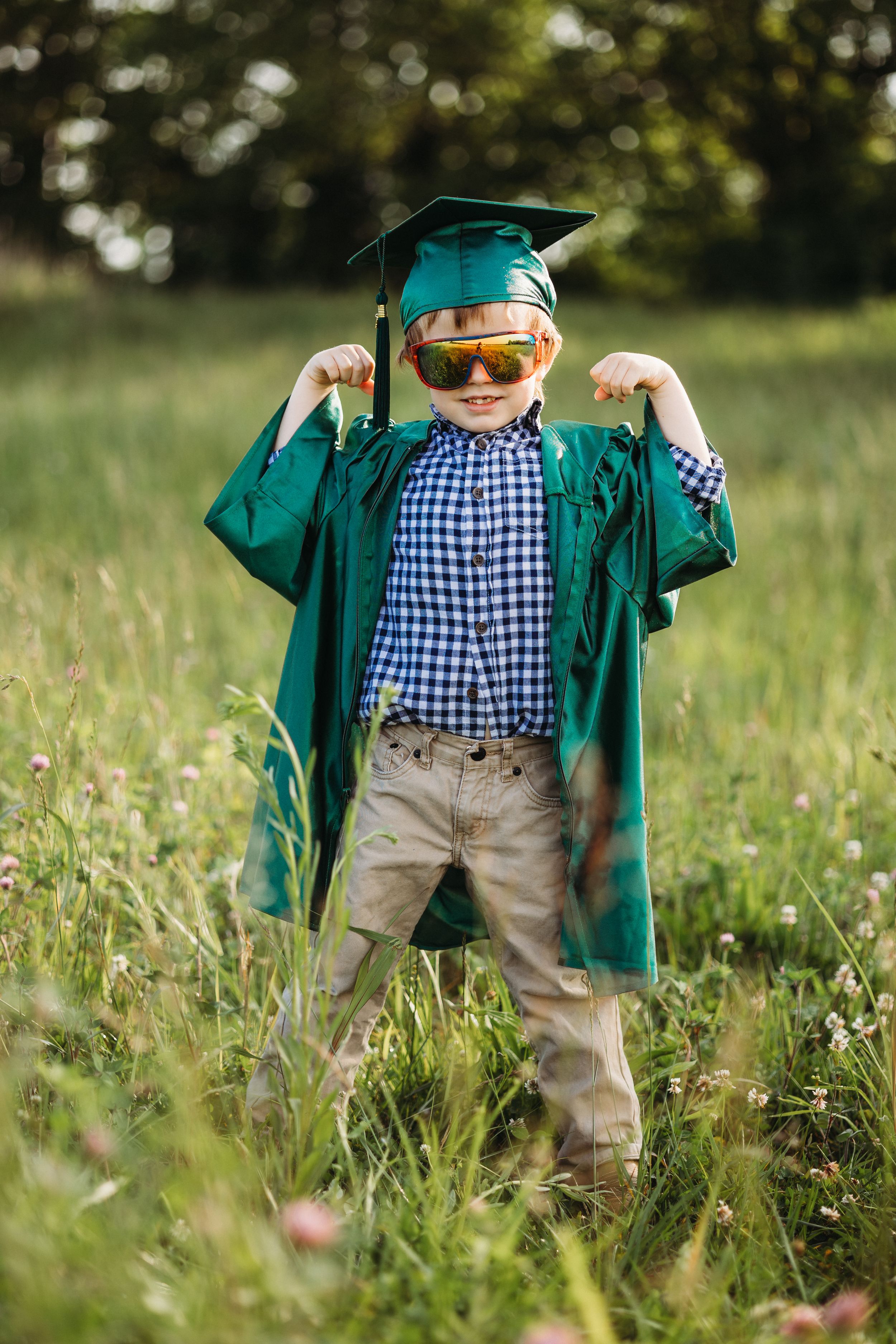 Young boy in green graduation cap and gown flexing muscles outdoors in a sunny field.