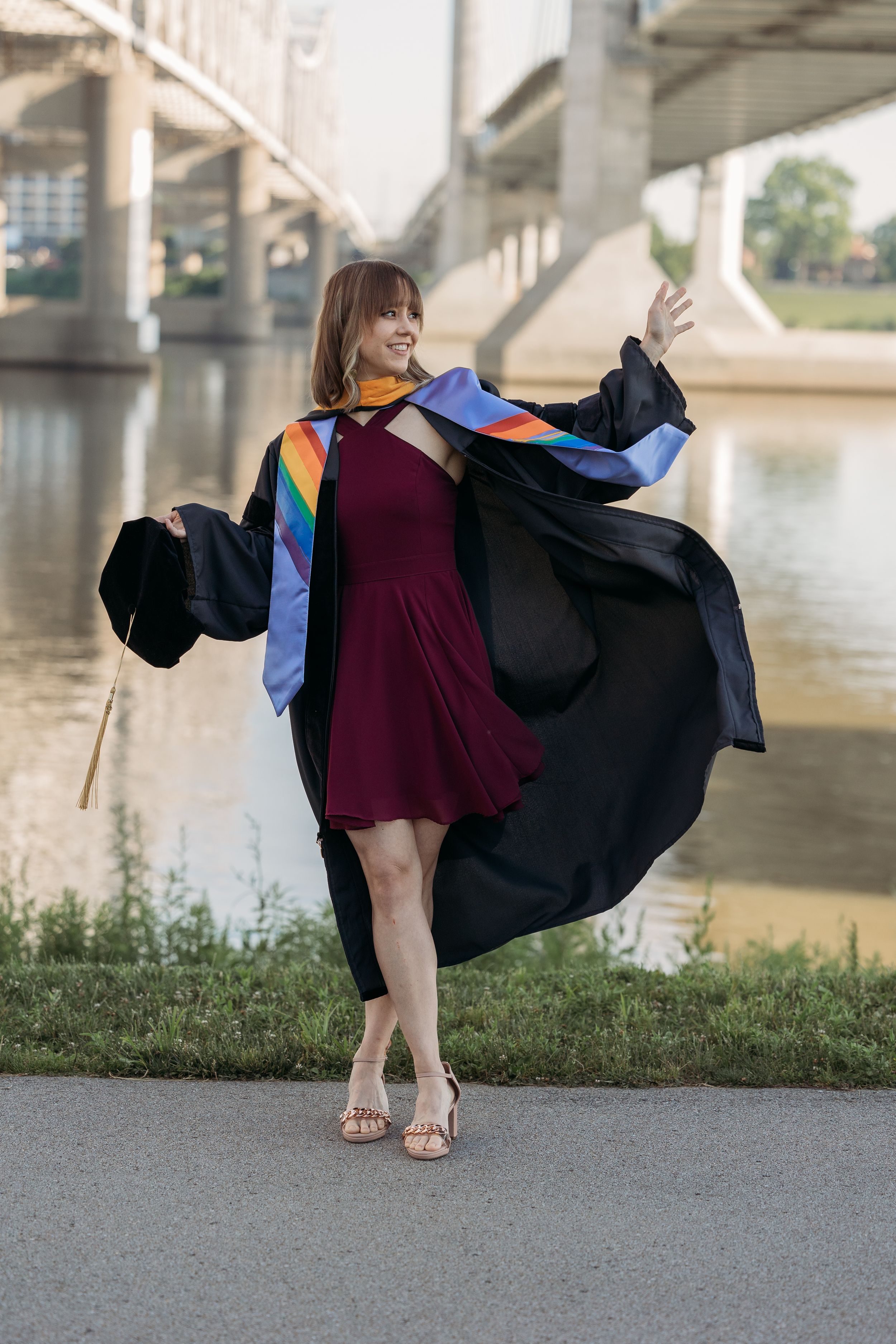 Woman in graduation gown with rainbow stole celebrating outdoors near a river and bridge.