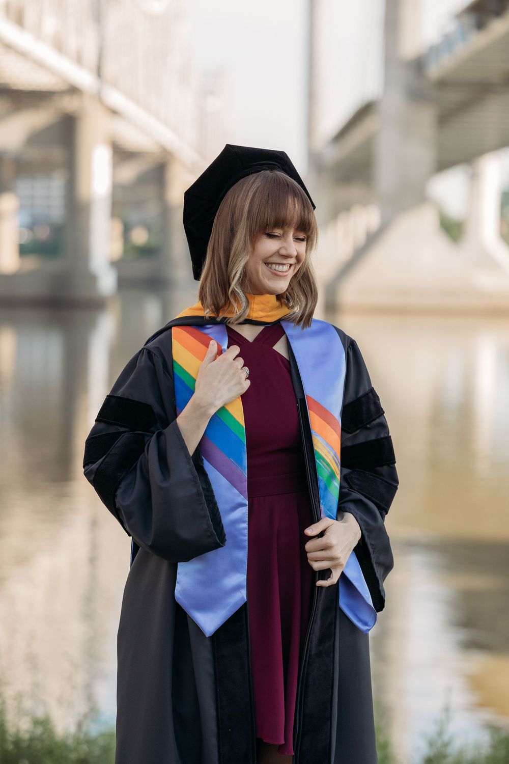 Smiling graduate in black doctoral gown and rainbow stole poses outdoors near a river bridge.