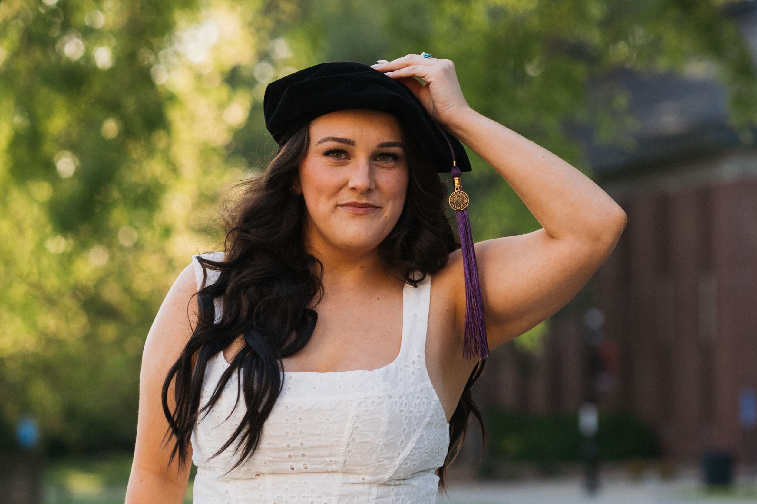 Young woman in white dress wearing graduation cap with purple tassel, smiling outdoors on college campus.