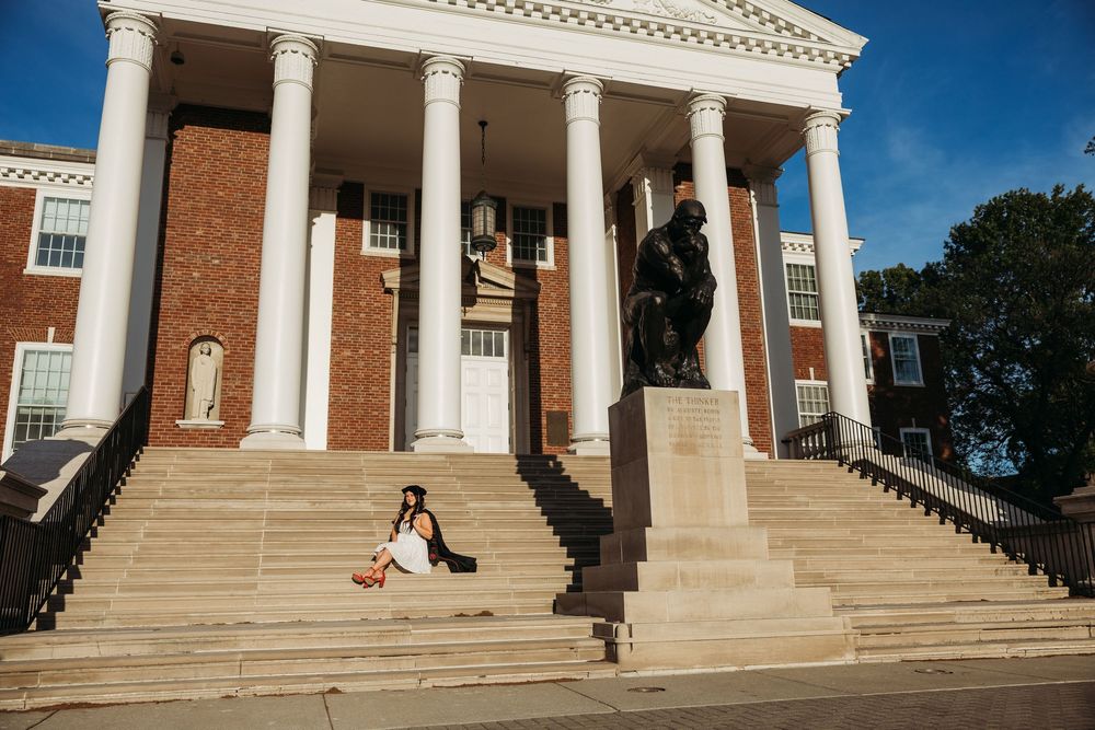 A person sits on the steps of a grand brick building with white columns and a bronze statue in the foreground.