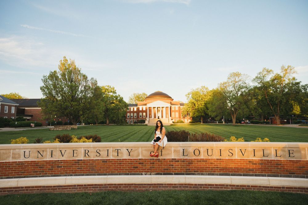 Couple posing in front of University of Louisville brick sign with Grawemeyer Hall dome and green campus lawn in background.