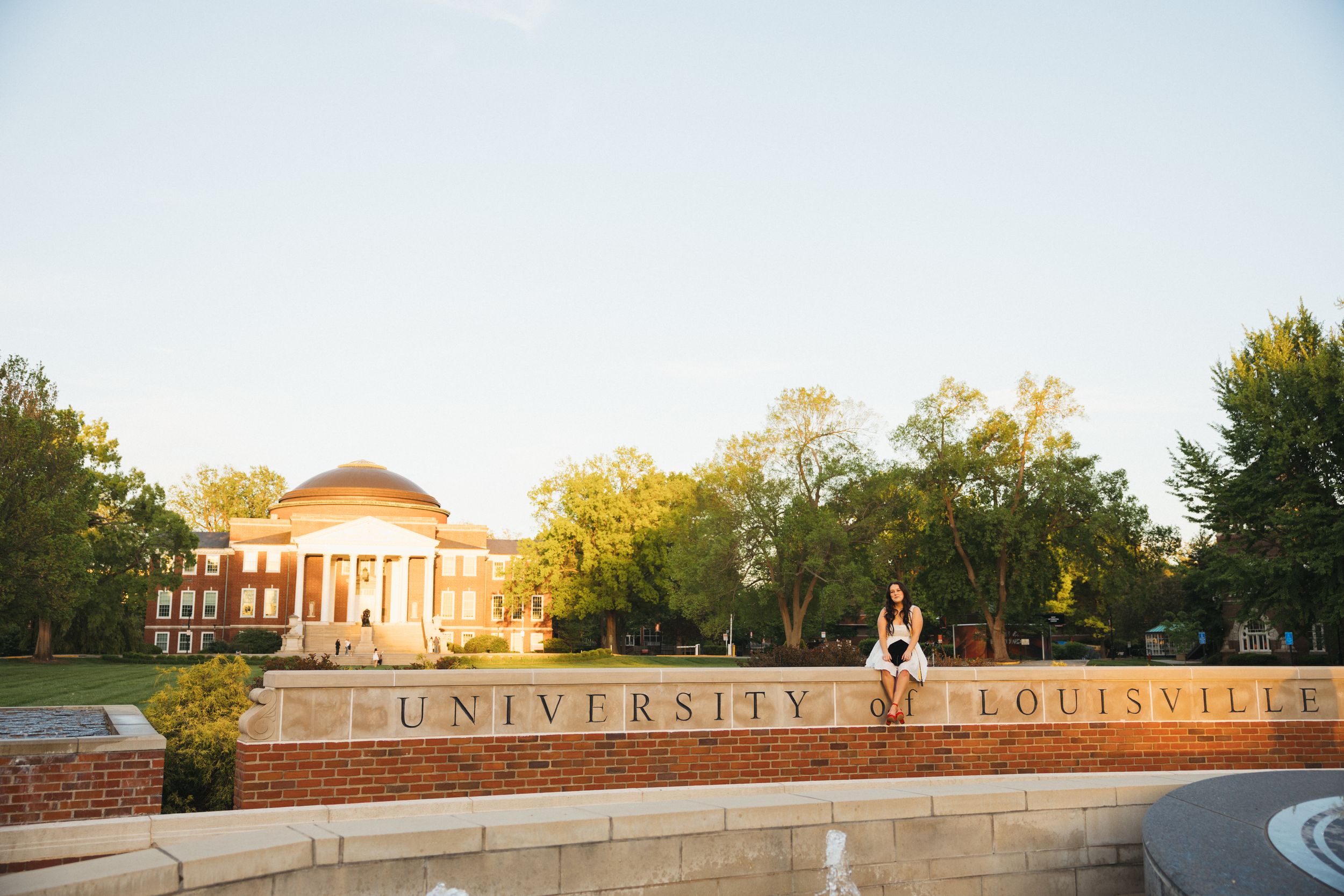 A student sits atop the University of Louisville brick sign with Grawemeyer Hall and lush trees in the background.