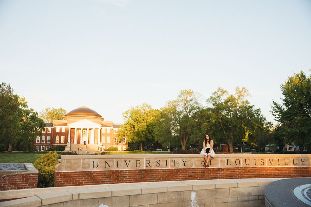 A student sits atop the University of Louisville brick sign with Grawemeyer Hall and lush trees in the background.