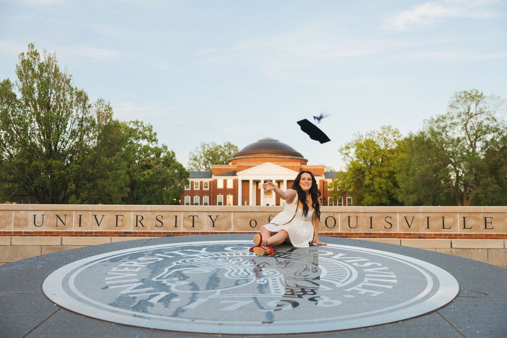 Woman sitting on University of Louisville seal, tossing graduation cap in air, with iconic campus building in background.