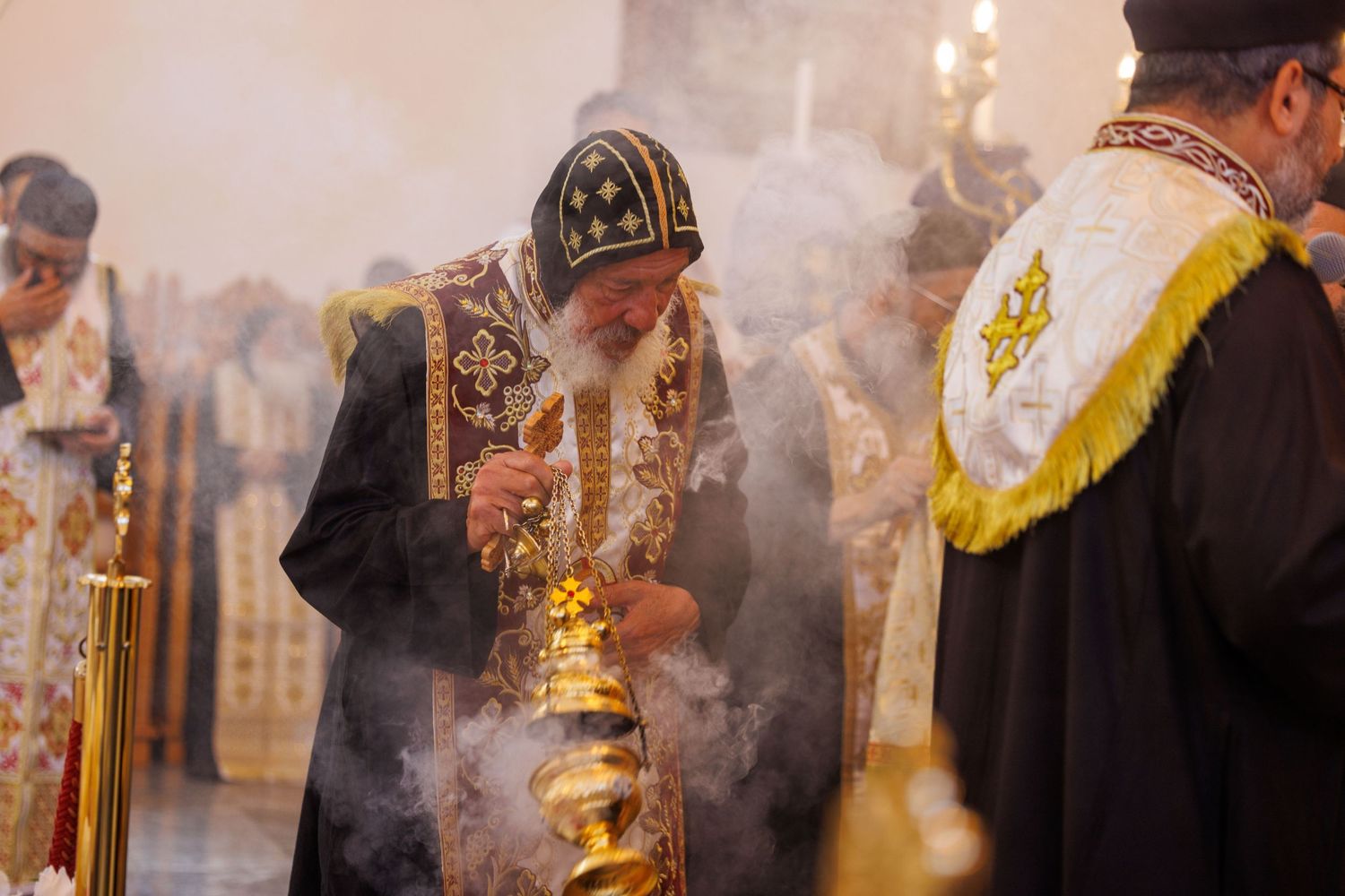 Orthodox bishop in ceremonial robes swinging a golden censer with incense smoke during a religious liturgy ceremony.