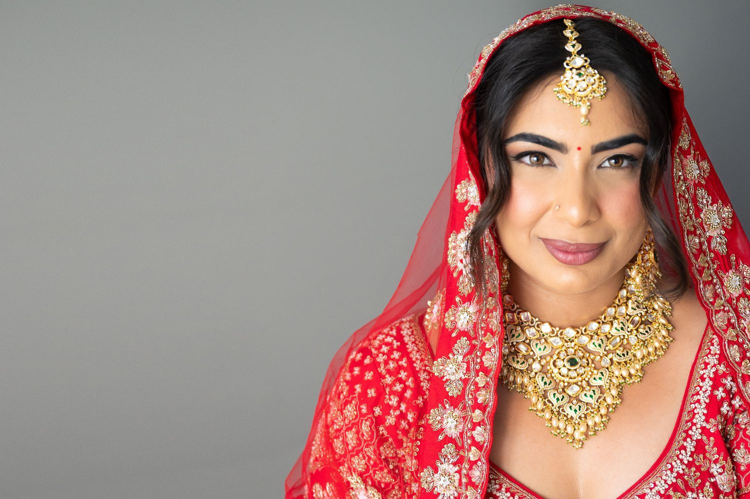 Indian bride in red lehenga and gold jewelry smiling, wearing traditional bridal dupatta and ornate necklace.