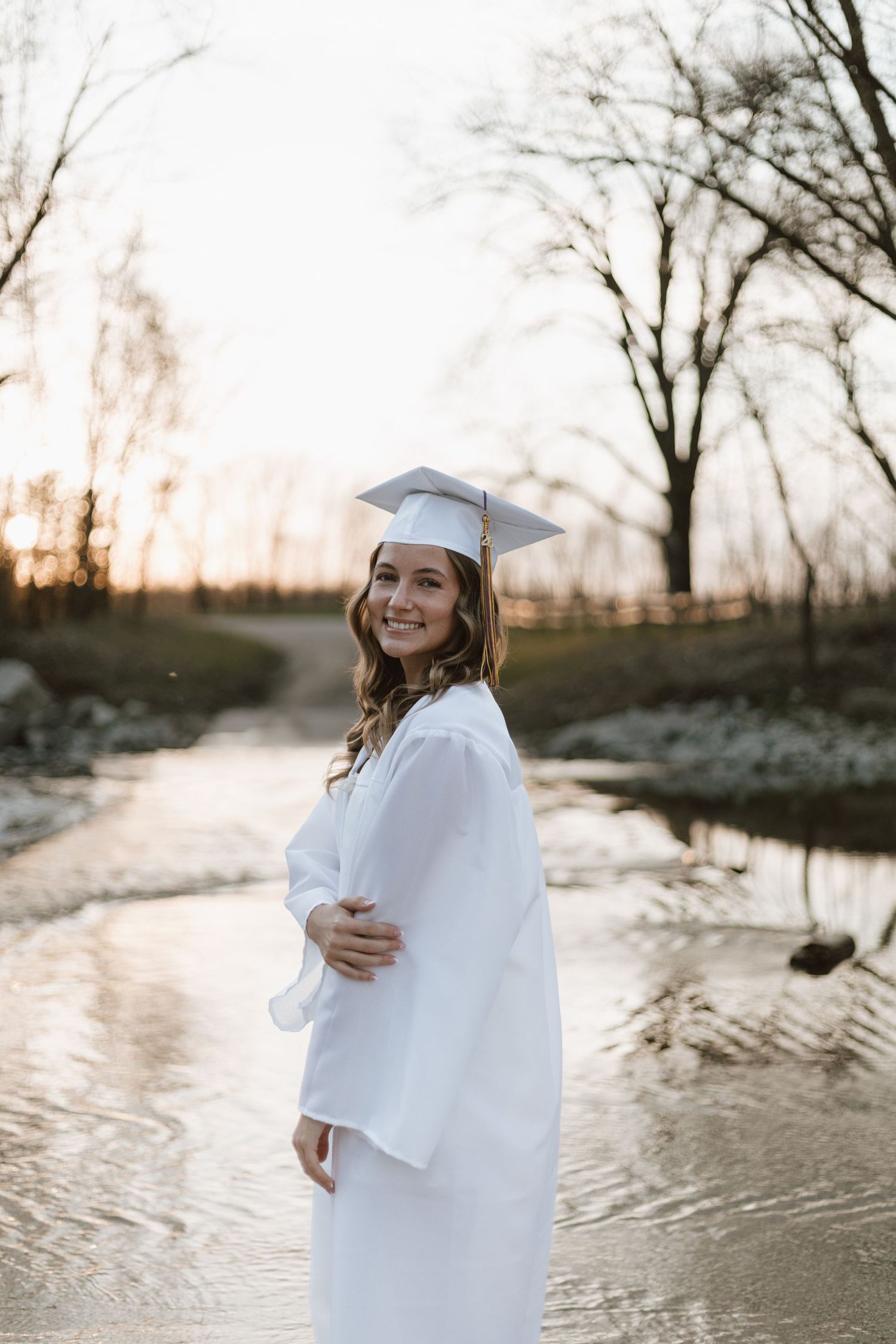 Young woman in white graduation cap and gown smiling outdoors by a creek at sunset.
