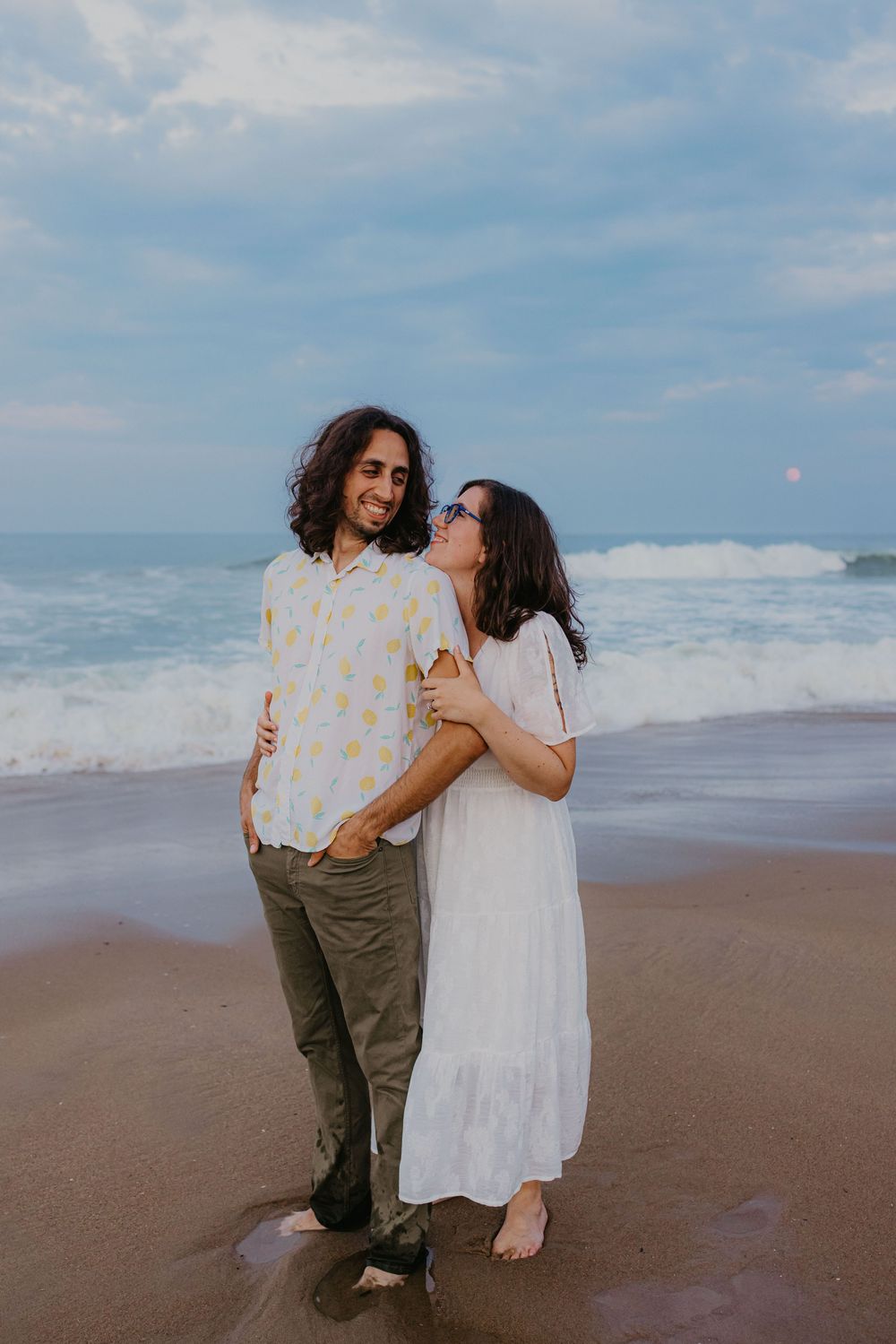 Joyful couple embraces barefoot on sandy beach with ocean waves crashing behind them at dusk.