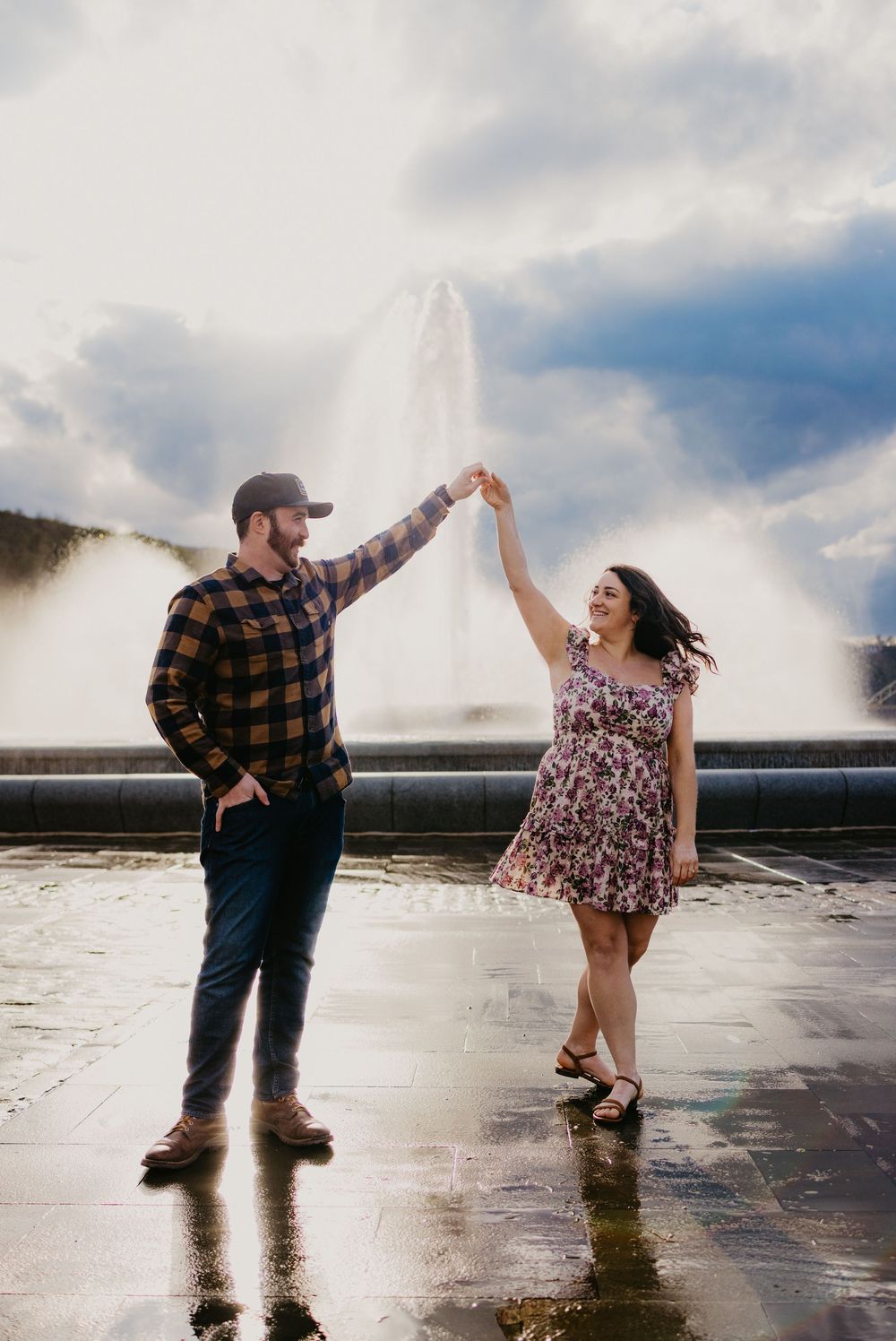 Couple dancing by a fountain, man in plaid shirt twirling woman in floral dress under dramatic cloudy sky.