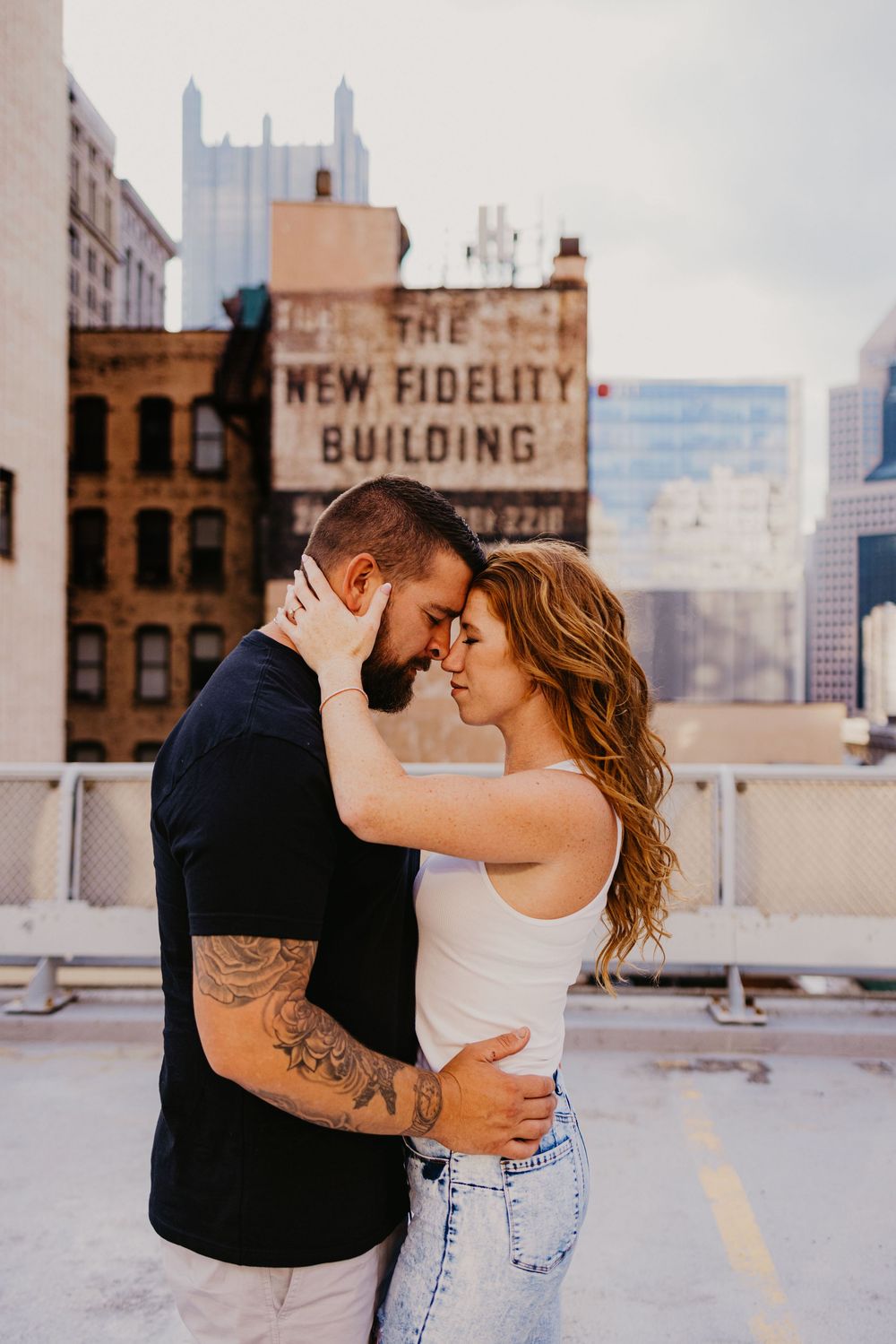 Couple embracing on rooftop with New Fidelity Building in Pittsburgh cityscape background, man with tattoos and woman in white tank.