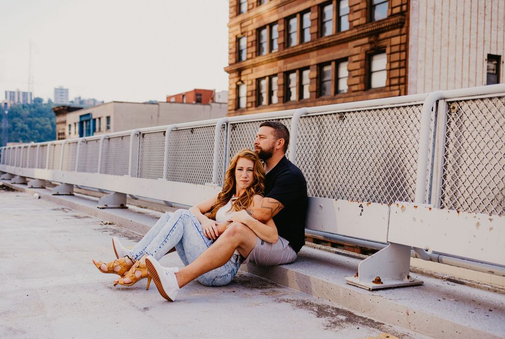 Couple sitting together on urban rooftop, leaning against metal railing with city buildings in background.