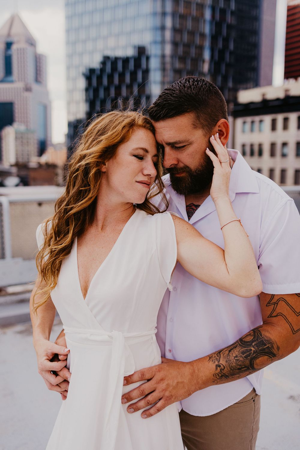 A couple shares a romantic moment on a rooftop with city skyline backdrop, woman in white dress, man with tattoos.