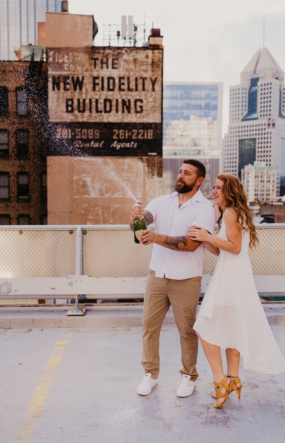 Couple celebrating on rooftop popping champagne with city skyline and New Fidelity Building backdrop.