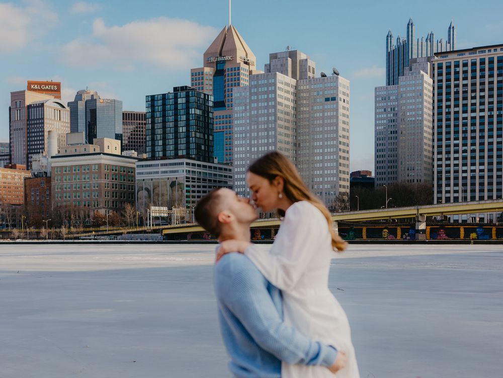 Couple sharing a romantic kiss on a frozen river with the Pittsburgh city skyline in the background.