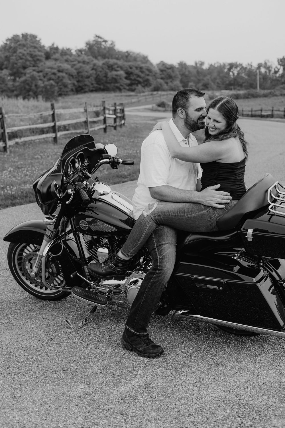 Black and white photo of a couple embracing on a Harley-Davidson motorcycle on a rural road.