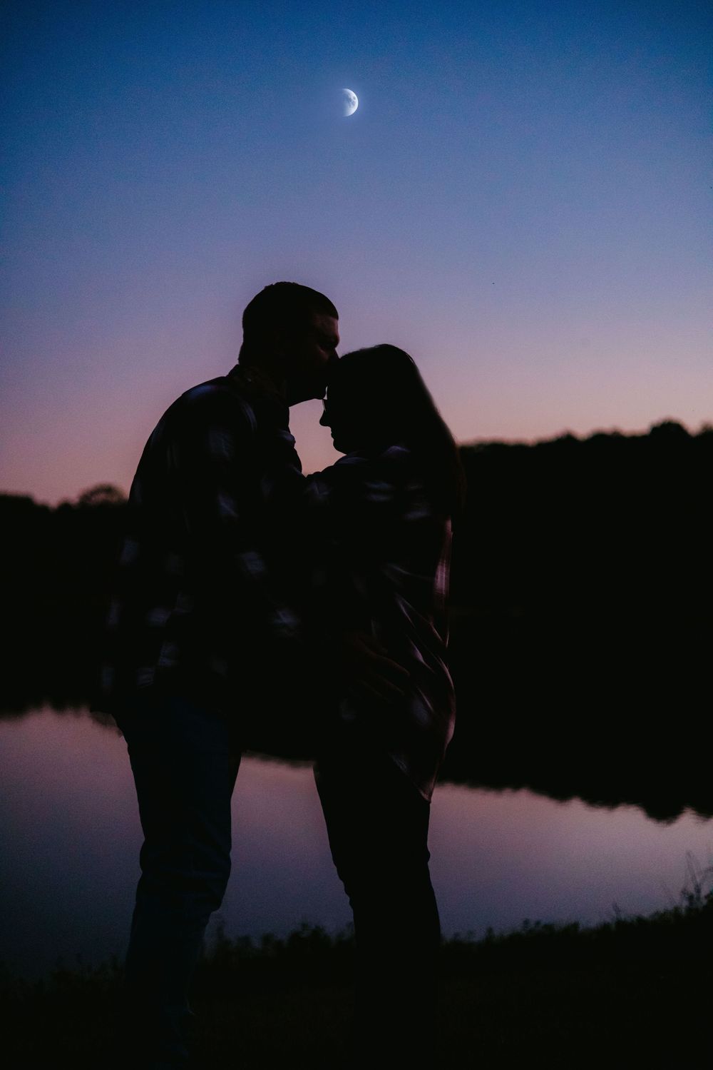Romantic couple silhouette sharing a tender moment by a reflective lake under a crescent moon at twilight.